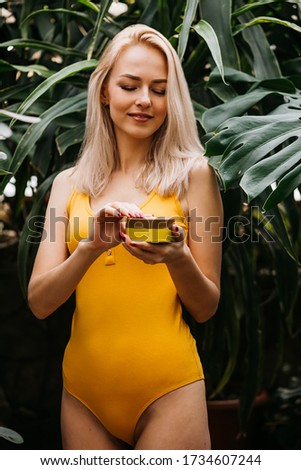 Beautiful caucasian blonde woman with healthy smooth clean skin holding jar of body cream, applying sunscreen, standing on tropical green leaves background. Concept of natural cosmetics and skin care.