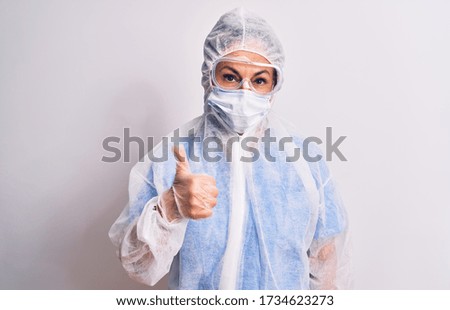 Middle age nurse woman wearing protection coronavirus equipment over white background doing happy thumbs up gesture with hand. Approving expression looking at the camera showing success.