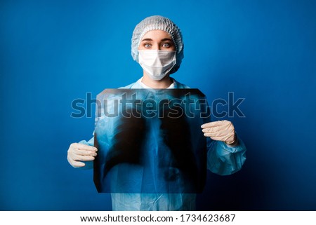Doctor renginologist in blue uniform standing on a blue background in a medical protective mask holding a chest x-ray in front of him, leaning a picture of his lungs against his chest