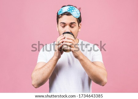 Portrait of nice calm peaceful sleepy young man with blue sleeping mask and cup of coffee or tea. Isolated over pink background.