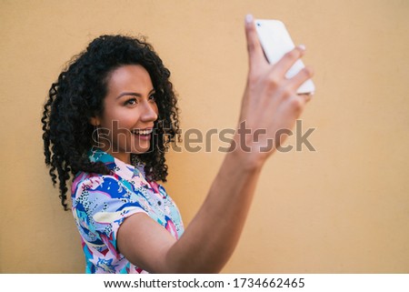 Portrait of afro woman taking selfies with her mophile phone against yellow wall. Technology concept.