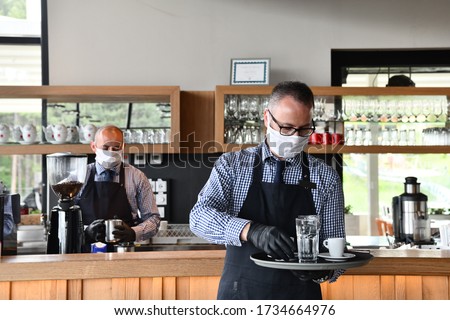 waiter in a medical protective mask serves  the coffee in restaurant durin coronavirus pandemic representing new normal concept Royalty-Free Stock Photo #1734664976