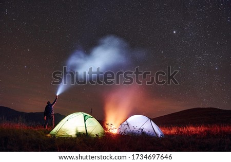 Man holds device with light beam. Two iluminated tents with campfire under stars at mountains at night.
