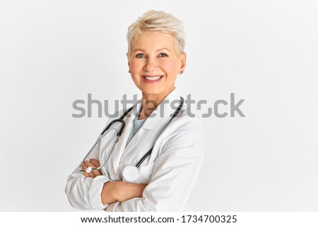 Beautiful successful mature woman pediatrician posing against white studio wall background with copy space for your advertisement, crossing arms on her chest, looking at camera with confident smile