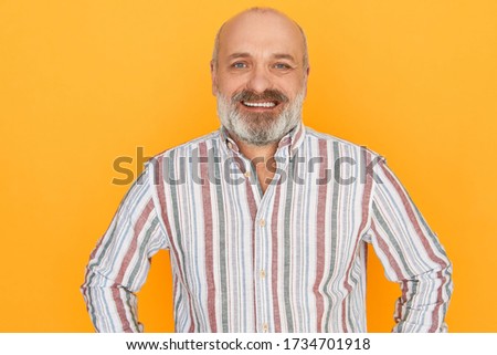 Positive human emotions and feelings. Picture of handsome bearded elderly male with carefree joyful smile enjoying good day, posing against yellow copy space wall background in striped shirt