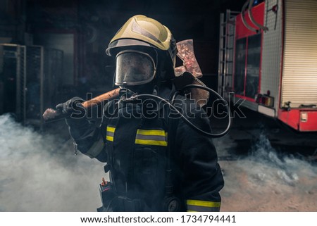 Portrait of a fireman wearing firefighter turnouts holding a rescue axe. Dark background with smoke and blue light.
