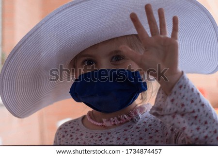 The little girl put on a medical mask, a white hat and a beads and looks out the window. Girl is ready to get out of isolation. The end of the Covid-19 pandemic