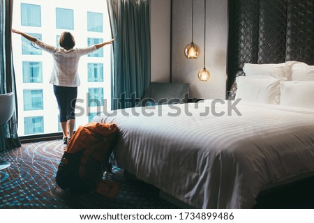 Backpacker female accomodating in 5 stars luxury hotel. She opening a whole wall window curtains to bring a day light into room. Solo traveling with backpack or home returning concept image. Royalty-Free Stock Photo #1734899486