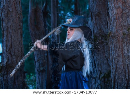 Woman in a witch costume in the forest at twilight holding a wooden stick