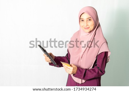 Portrait photo of Muslim woman, age 30-35, wearing pink hijab, dark pink cloth holding tablet and credit card isolated on white background, concept of online shopping and payment.