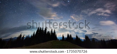 Panoramic view of a mountain hill facing spruce forest and a mountain ridge, magical starry night sky with purple clouds above beautiful horizon, copy space. Concept of travelling, galaxy and nature.