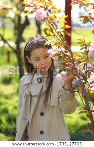A young beautiful girl in a beige cloak and pants stands near a flowering tree.
The photo was taken with selective focus.
