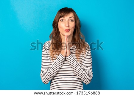 Portrait of young friendly woman with essence in a casual t-shirt makes prayer gesture on an isolated blue background. Emotional face. To ask for something from the universe or from God, make a wish