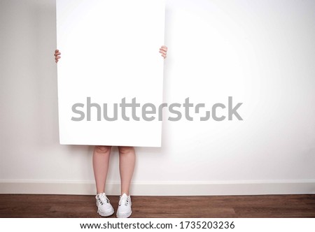 Young woman holding a blank poster, board or frame for text against white wall in modern room interior space for text