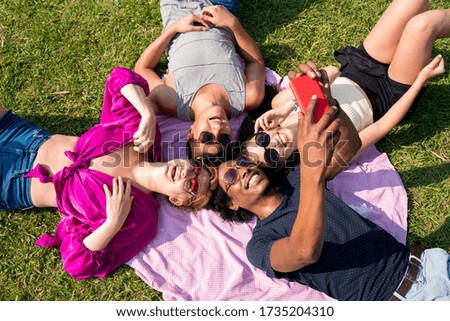 A group of multiethnic friends taking selfies while laying in the grass on a sunny day in a park.