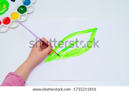 Top view of child is drawing green leaf by watercolors on white paper. Earth day and environmental protection concept.