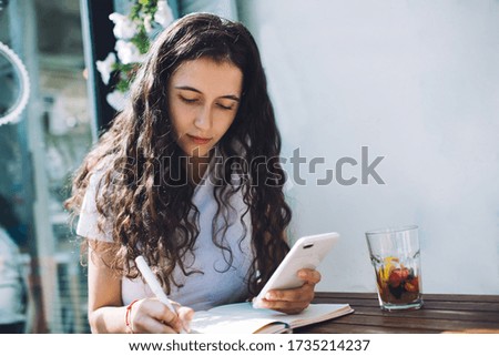 Serious young female with long curly hair in white tee shirt using mobile phone and taking notes in notebook while sitting in outdoor cafe