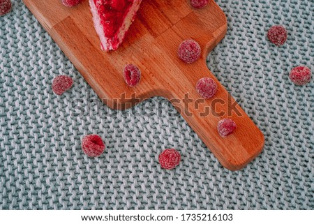  A raspberry cheesecake on a wooden chopping Board lies on a blue plaid of large knitting, frozen raspberries are scattered everywhere