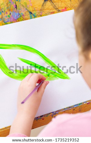 Close up of hand of child is drawing green leaf with brush by watercolors on white paper on an easel. Earth day concept.