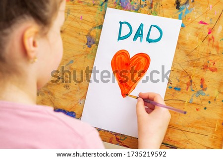 Back view of littel girl is drawing red heart with word Dad greeting card by watercolors on white paper on an easel. Father's Day concept.