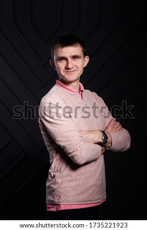 Young brunette man, wearing casual light pink jumper, posing for picture in front of black wall in photo studio, smiling, laughing, with arms crossed on chest. Corporate culture. Happy Emotions.