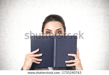 Portrait of young businesswoman or student with dark hair covering her face with blue book near concrete wall. Concept of knowledge and education