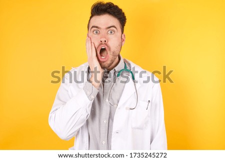 Horizontal portrait of shocked handsome caucasian doctor man wearing medical uniform looks with great surprisment being very stunnedwith unexpected news, models against yellow background. 