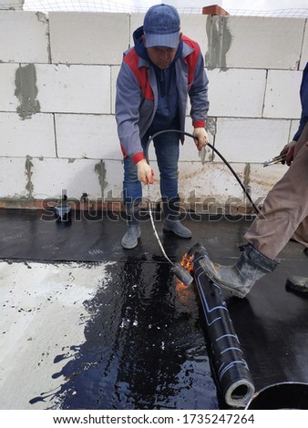 A waterproofing specialist with an assistant melts the bituminous resin with a blowtorch and seals a reliable roll material. Hermetic covering of roofs and walls at a waterproof. Royalty-Free Stock Photo #1735247264