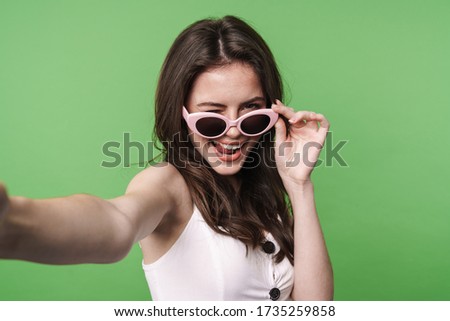 Portrait of a beautiful excited young brunette girl with long hair wearing summer dress standing isolated over green background, taking a selfie in sunglasses, winking