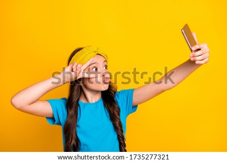 Photo of funny little lady hold telephone quarantine time stay home talk skype classmates show v-sign near eye send kisses wear casual blue t-shirt headband isolated yellow color background