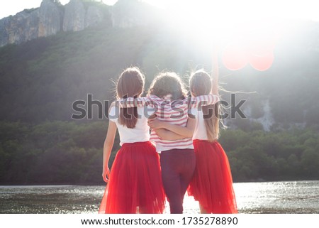 Back view of group of pretty girls dressed red and white clothes, sunglasses walking with fun in the nature together and holding the balloons. Preteen people playing against the beautiful view 