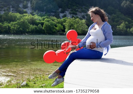 Adult pregnant woman relaxing in the nature against the lake . Pretty girl looking to the tummy  with love, female dressed blue jeans, white t-shirt, and shirt, holding red  baloons