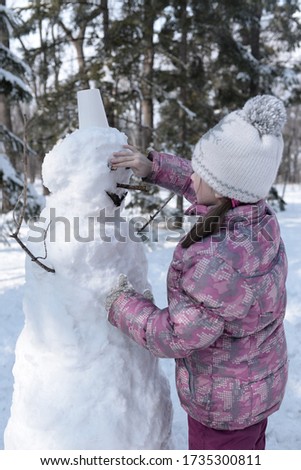 Girl sculpts a snowman. Winter forest. Trees in the park in the snow. Girl in pink overalls and a white hat.