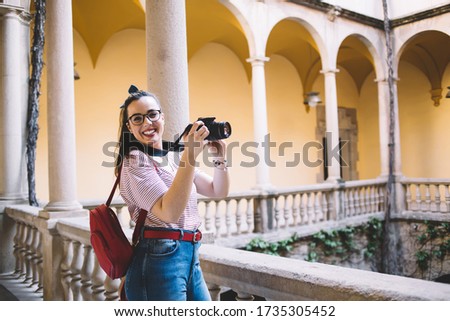 Portrait of cheerful caucasian female photographer enjoying work process using modern equipment outdoors, smiling woman tourist holding digital camera taking amateurs photo during free time