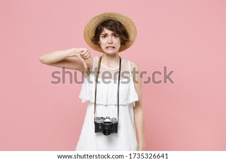 Confused displeased young tourist woman in summer dress hat with photo camera isolated on pink background. Female traveling to travel weekend getaway. Air flight journey concept. Showing thumb down