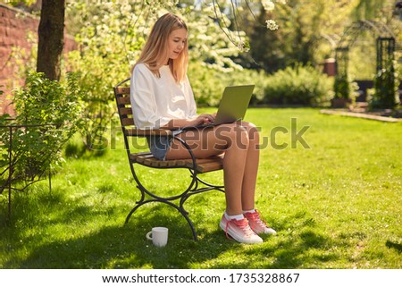 A cute teen girl is texting on social networks on a laptop, sitting on a chair in the yard, on the lawn, in the garden.