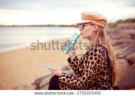 Beautiful blonde woman wearing leopard print coat and a hat resting on the beach. She is drinking watter from a bottle and using mobile phone to take pictures.