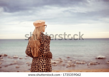 Beautiful young blonde woman looking on the sea, she is resting on the beach. She is wearing leopard print coat and a hat. 