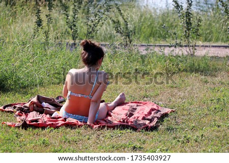 Lonely woman in a swimsuit sitting with smartphone on the grass by the river. Beach holidays and sunbathing in a park during coronavirus pandemic