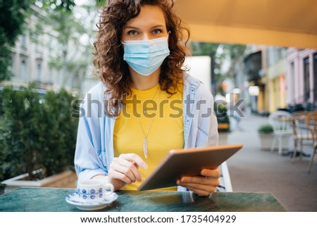Portrait of young woman wearing protective medical mask sitting in street cafe holding digital tablet in her hands at summer day.