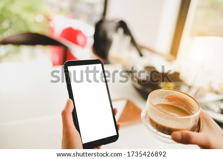 Mockup image of hand’s woman holding black mobile phone with blank screen and hot latte coffee on vintage wood table in cafe.