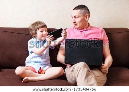 Surprised young man sit on computer using laptop relax with preschooler son have fun together, dad and little boy child enjoy smartphone at home rest on sofa busy with gadgets. Online education.