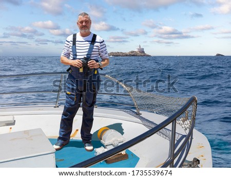 The captain of a ship stands at the bow with binoculars and looks into the camera. In the background the small Spanish rocky island Isla Hormiga, which translates as an ant island.