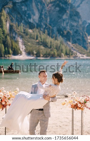 The groom circles the bride in her arms at the venue for ceremony, with two autumn flower columns instead of an arch, against backdrop of Lago di Braies in Italy. Wedding in Europe, on Braies lake.
