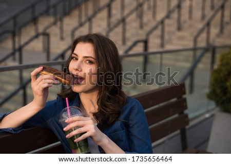young beautiful woman going to bite cake on city street, brunette girl in blue shirt and blue jeans sitting on bench
