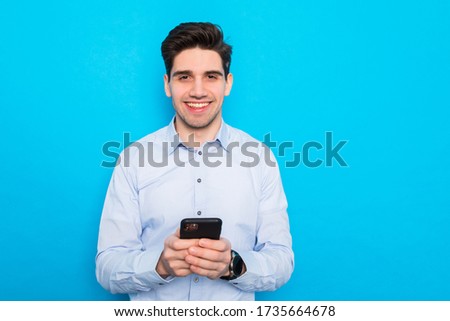 Smiling young man holding mobile phone, looking at screen isolated on blue studio background.