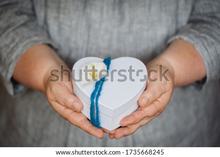 Hands holding a small white gift box in heart shape with a self-made blue, turquoise gift ribbon and a flower. shallow depth of field