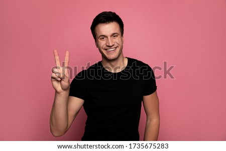 Peace for all. Close-up photo of a charismatic young man in a black t-shirt, who is looking in the camera with a big smile and showing a peace sign.
