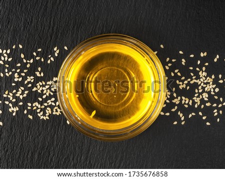 Sesame seed oil in a glass bowl and white sesame seeds on a black stone background, flat lay. Useful natural oil.