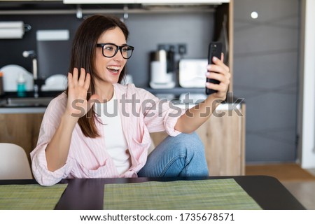 Smiling woman sitting on kitchen sofa talking by videocall dating online looking at phone. Video blogger vlogger recording vlog at home. Lifestyle vlogging concept, head shot portrait Royalty-Free Stock Photo #1735678571